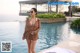 A woman standing next to a swimming pool wearing a brown dress.