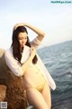 A woman in a white shirt and a yellow bikini posing by the ocean.