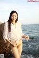 A woman in a white shirt and gold bikini standing on a rock by the ocean.