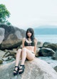 A woman in a black bikini sitting on a rock by the water.